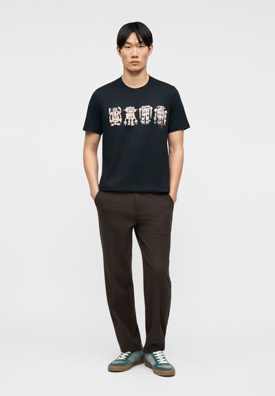 Άνδρας που φοράει μαύρο μπλουζάκι με τέσσερις ροζ εικόνες ρομπότ, σκούρο καφέ παντελόνι, πράσινα αθλητικά παπούτσια, στέκεται με τα χέρια στις τσέπες σε λευκό φόντο.