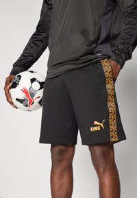 Shorts de sport noirs avec le texte orange "KING" et des bandes latérales à motifs. Le modèle tient un ballon de football noir et blanc.