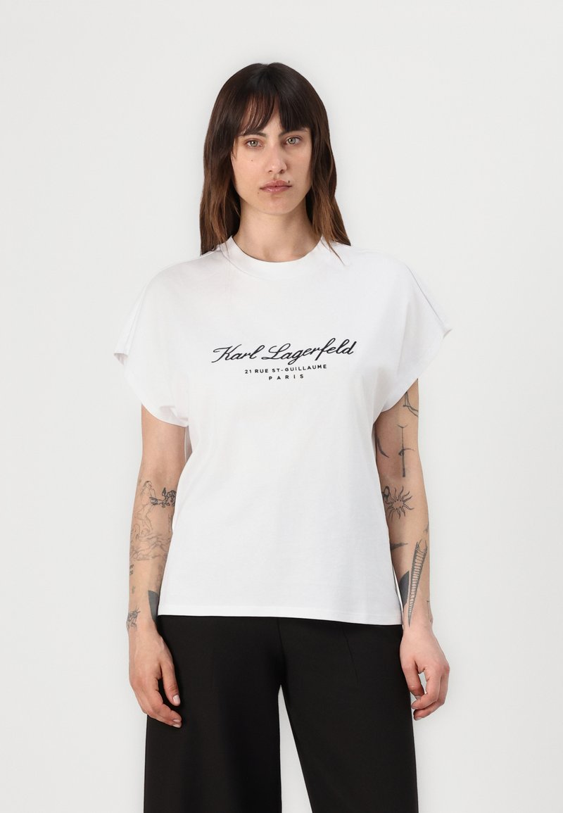 White cotton t-shirt with short sleeves and a crew neckline. Features black print: "Karl Lagerfeld 21 RUE ST. GUILLAUME PARIS" on the front.