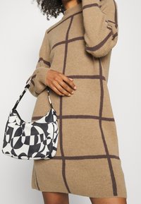 Una persona con capelli ricci indossa un maglione color cammello con motivi a quadri marroni, tenendo una borsa a tracolla con motivo bianco e nero.