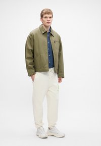 Olivovo zelená bunda cez denimovú košeľu, doplnená krémovými teplákmi s neónovo zeleným logom. Bielym teniskám je outfit dokončený.