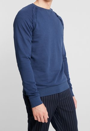 Mann trägt ein eng anliegendes, langärmeliges Marineblaues Sweatshirt und schwarze Nadelstreifenhose, steht vor einem schlichten hellen Hintergrund.