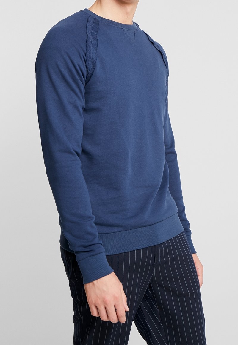 Homme portant un sweat-shirt bleu marine à manches longues ajusté et un pantalon noir à fines rayures, debout devant un fond clair uni.