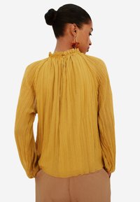 Blusa gialla plissettata con scollo alto, ruffles arricciati e maniche lunghe, dotata di chiusura con bottoni sul retro.