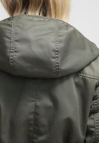 Olivově zelená bunda s kapucí vyrobená z hladké látky, s jednoduchým designem viditelným švem na švu a uvolněným střihem.