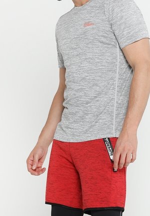 Man draagt een nauwsluitend grijs shirt met korte mouwen en rode sportshorts met een zwart-wit "Superdry" label aan de zijkant.
