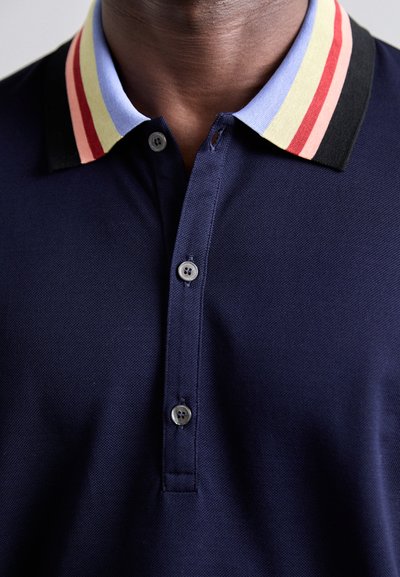 T-shirt polo bleu marine en tissu texturé, avec un col rayé multicolore en noir, rouge, jaune et bleu. Patte de boutonnage à deux boutons.