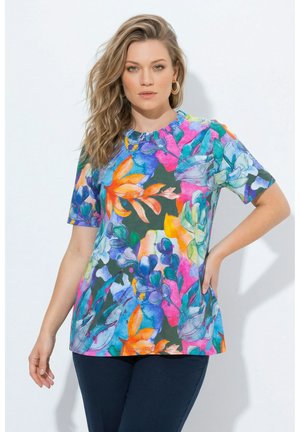 Femme aux cheveux blonds ondulés, portant un haut coloré à manches courtes à motifs floraux et un pantalon foncé, debout avec une main sur la hanche.