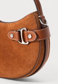 Brūnas zamšādas rokas soma ar izliekta formu, ar ādas siksnu un sudraba toņa aparatūras akcentiem. Pa malām ir redzama detalizēta dūriena šuvēm.