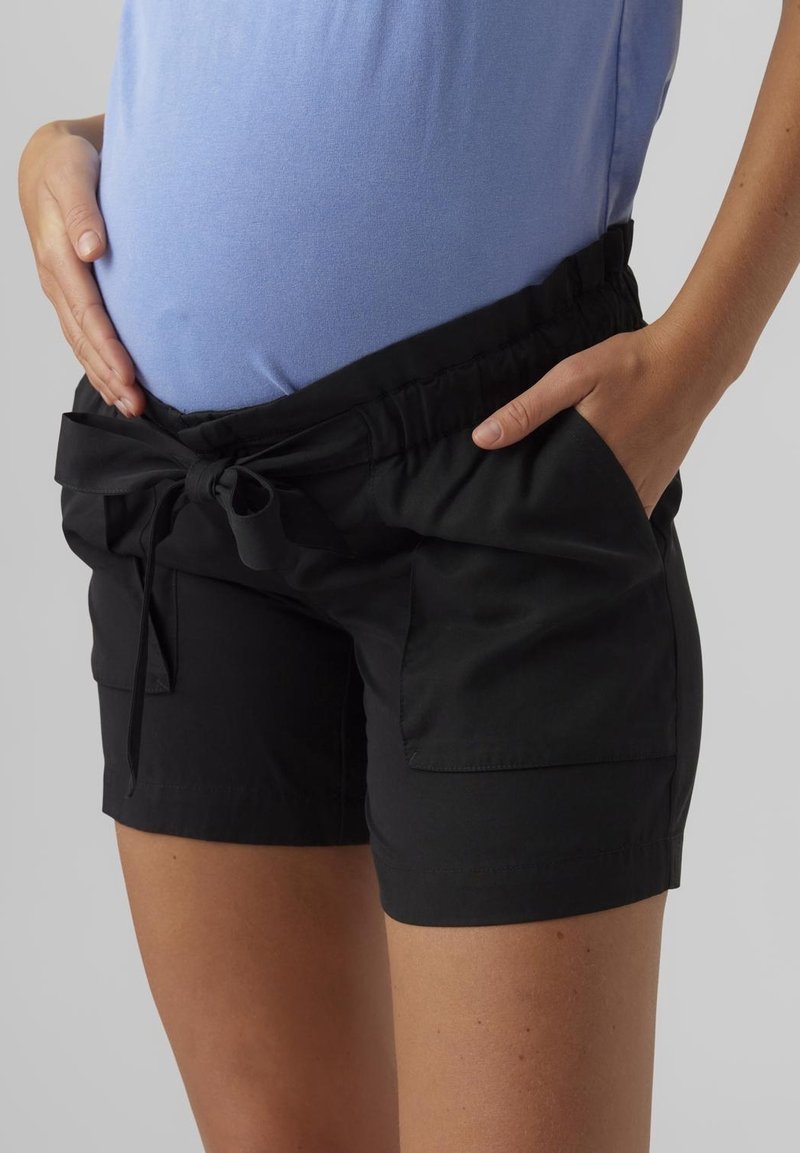 Raska gravid med svarta gravidshorts med framknutning, ena handen i fickan och den andra vilande på magen.