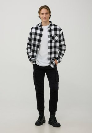 Camicia button-up in motivo a quadri bianco e nero, abbinata a pantaloni cargo e stivali combat neri. Il materiale sembra robusto con una vestibilità casual.