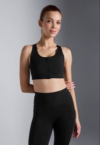 Sujetador deportivo negro con cremallera frontal y tirantes anchos, fabricado con una tela elástica. Combinado con leggings negros de tiro alto que cuentan con una cinturilla acanalada.