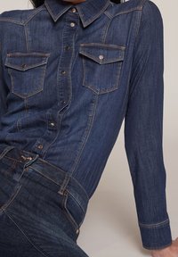 Denim shirt in dark blue with a pointed collar, two chest pockets, and contrasting orange stitching along the seams and buttons.