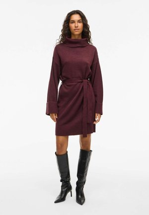 Robe pull bordeaux avec un col montant, des manches longues et une taille ceinturée. Comprend des poignets côtelés et un tissu lisse et texturé. Bottes noires jusqu'aux genoux.
