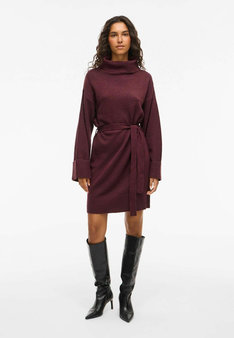 Robe pull bordeaux avec un col montant, des manches longues et une taille ceinturée. Comprend des poignets côtelés et un tissu lisse et texturé. Bottes noires jusqu'aux genoux.