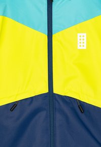 Elölről nézve színes blokkokból készült dzseki, amely tealkék, sárga és sötétkék panelekből áll, középen zippzárral és két fekete zippzárhúzóval.