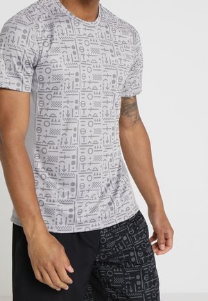 Homme portant un t-shirt blanc à manches courtes et un short noir, tous deux ornés de motifs géométriques composés de lignes, cercles et triangles.