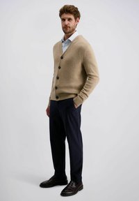 Beige cardigan met V-hals, zes zwarte knopen en geribde manchetten, gecombineerd met donkerblauwe broek en zwarte schoenen, staand op een neutrale achtergrond.