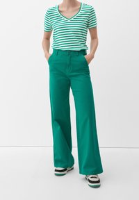 Zelené kalhoty s širokými nohavicemi v kombinaci se zeleným a bílým pruhovaným tričkem s výstřihem do V. Obsahují přední kapsu a kontrastní švy.