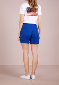 Kvinna med långt hår står vänd bort med vita sneakers, blå uppvridna shorts och en vit t-shirt med en amerikansk flagga söm på ryggen.