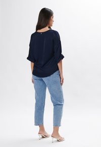 Marineblaue, locker sitzende Bluse mit umgeschlagenen Ärmeln und einem Schlüssellochdetail auf der Rückseite, kombiniert mit hellblauen High-Waist-Jeans und weißen hochhakigen Sandalen.