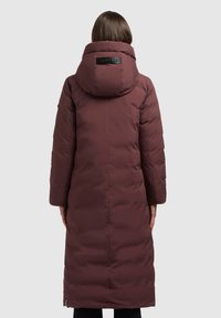 Długi pikowany płaszcz w kolorze burgundowym, z dużym kapturem, minimalistycznym wzornictwem i naszywką z logo z tyłu. Gładka, izolowana tekstura.