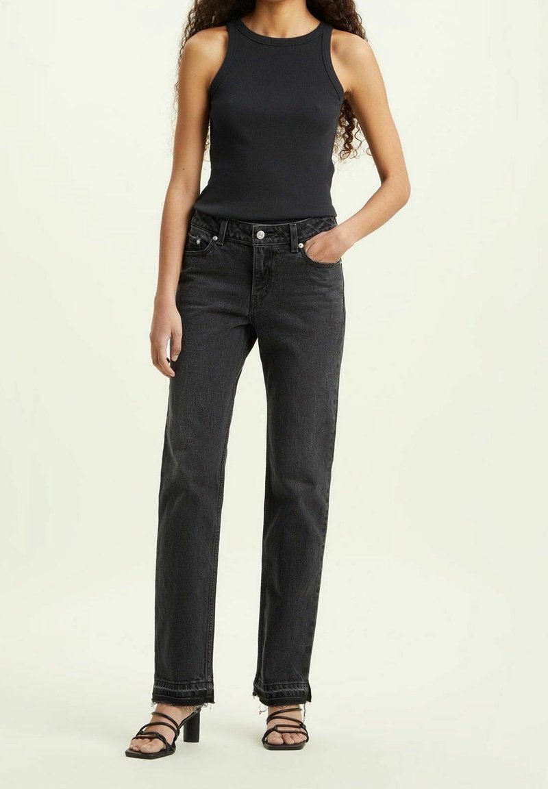 Jeans de mezclilla negra con corte recto, cintura alta y dobladillo desgastado, combinados con un top negro sin mangas ajustado y tacones con tiras.