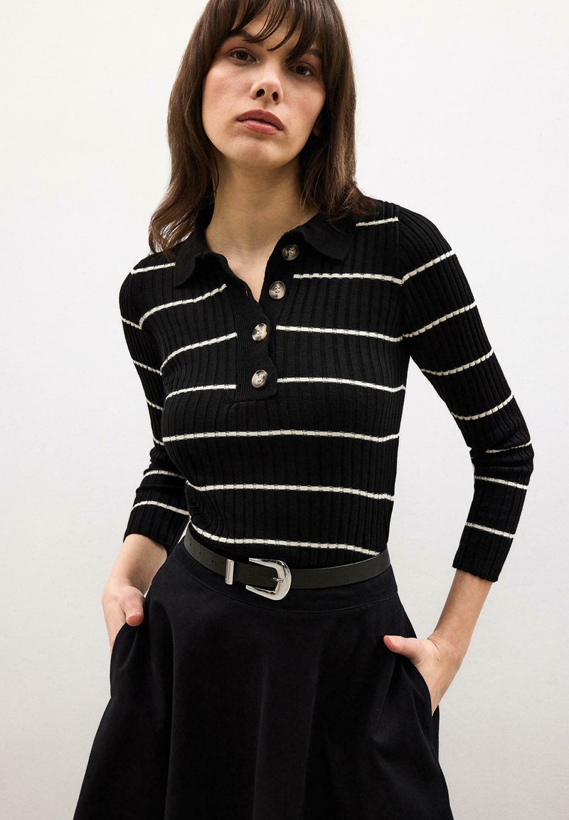 Femme portant un polo noir côtelé à manches longues avec des rayures blanches et quatre boutons, une jupe noire et une ceinture noire avec une boucle argentée, mains dans les poches.