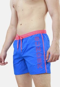 Pantaloni da nuoto blu con accenti rosa e cordino. Presenta "SUNDEK" in lettere in grassetto sul lato. Materiale leggero e liscio.