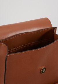 Ruskea nahkainen käsilaukku, jossa on sileä pinta, läpällinen yläosa ja kultainen nepparikiinnitys. Sisäosassa on järjestelyosasto.