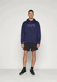 Άνδρας ντυμένος με μπλε φούτερ με καπάκι που φέρει το λογότυπο TYR, μαύρα αθλητικά σορτς, μαύρες κάλτσες και μαύρα αθλητικά παπούτσια σε απλό φόντο.