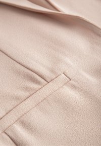 Detailní záběr na jemnou, světle béžovou texturu látky se dekoračním vertikálním švem a záhybem, který naznačuje, že se jedná o látku na šité oblečení.