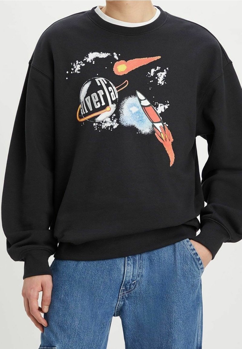 Sweat-shirt noir avec un design coloré de l’espace montrant une fusée, une planète avec des anneaux, une comète et des amas d’étoiles, porté avec un jean bleu.