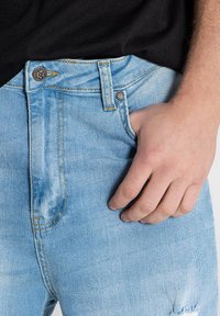 Jeans de mezclilla azul claro con desgaste, costuras visibles y un bolsillo frontal. La cinturilla cuenta con un botón de metal y detalles de bucle.