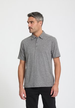 Man met grijs haar, gekleed in een grijs polo-shirt met korte mouwen en een zwarte broek, staand tegen een effen witte achtergrond.