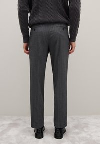 Graue Nadelstreifenhose mit maßgeschneiderter Passform, ausgestattet mit Gesäßtaschen und einem glatten, strukturierten Stoff. Kombiniert mit schwarzen Anzugschuhen.