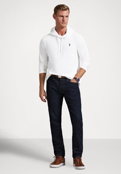 Fehér, hosszú ujjú kapucnis pulóver húzózsinórral, sötétkék farmer és barna sportcipő, barna övvel és kék órával viselve.