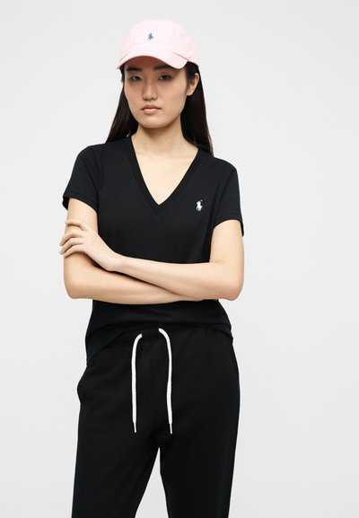 Γυναίκα με μακριά σκούρα μαλλιά, που φοράει ροζ καπέλο, μαύρο T-shirt με V λαιμόκοψη και μαύρο παντελόνι με λευκό κορδόνι, με σταυρωμένα τα χέρια.