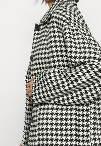 Płaszcz w czarno-białą kratkę houndstooth, wykonany z teksturowanej tkaniny, z dużymi czarnymi guzikami i kołnierzowym dekoltem.