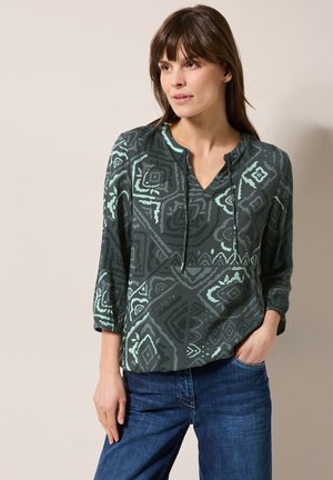 Blouse à motifs vert foncé avec un décolleté en V et des manches 3/4, présentant un design géométrique avec des accents vert clair et une coupe décontractée.
