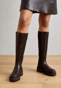 Botas de cuero marrón hasta la rodilla con suela gruesa y textura suave, que presentan un diseño sin costuras y mínimos acentos metálicos.