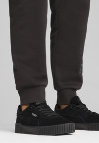 Schwarze Wildleder-Sneaker mit einer strukturierten Sohle, dezentem Branding und einem flachen Design, kombiniert mit dunklen schmal zulaufenden Jogginghosen.