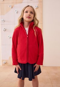 Giacca in pile rossa con collo alto, chiusura a zip e tasche laterali, indossata sopra una camicia bianca e una gonna blu navy. Tessuto liscio, design casual.