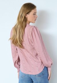 Femeie cu păr blond lung, purtând o bluză roz texturată cu detalii din dantelă și blugi albaștri, stând lateral lângă un perete alb.