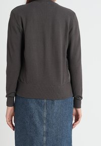 Persona vestida con una chaqueta de manga larga gris oscuro y una falda de mezclilla azul, de pie mirando hacia atrás contra un fondo claro y sencillo.