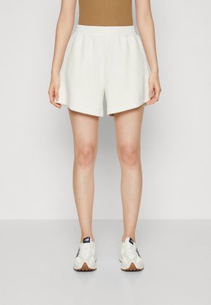 Personne portant un short blanc taille élastique, un top marron et des baskets blanches, debout devant un fond clair uni.