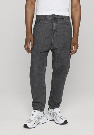 Personne portant un jean large gris foncé et des baskets blanches New Balance sur un fond clair uni.