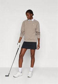 Kvinna i beige sweatshirt, svart kjol, vit golfhandske och sneakers som håller i en golfklubba, står mot en ljus enfärgad bakgrund.