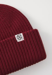 Beanie a coste bordeaux con risvolto, caratterizzato da un'etichetta quadrata bianca che mostra un logo circolare nero su sfondo bianco.