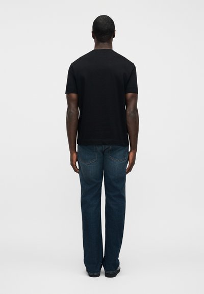 Μαύρο βαμβακερό t-shirt με στρογγυλή λαιμόκοψη, κοντά μανίκια, σε συνδυασμό με μπλε τζιν παντελόνι με τσέπες στο πίσω μέρος και ίσια γραμμή στο πόδι.
